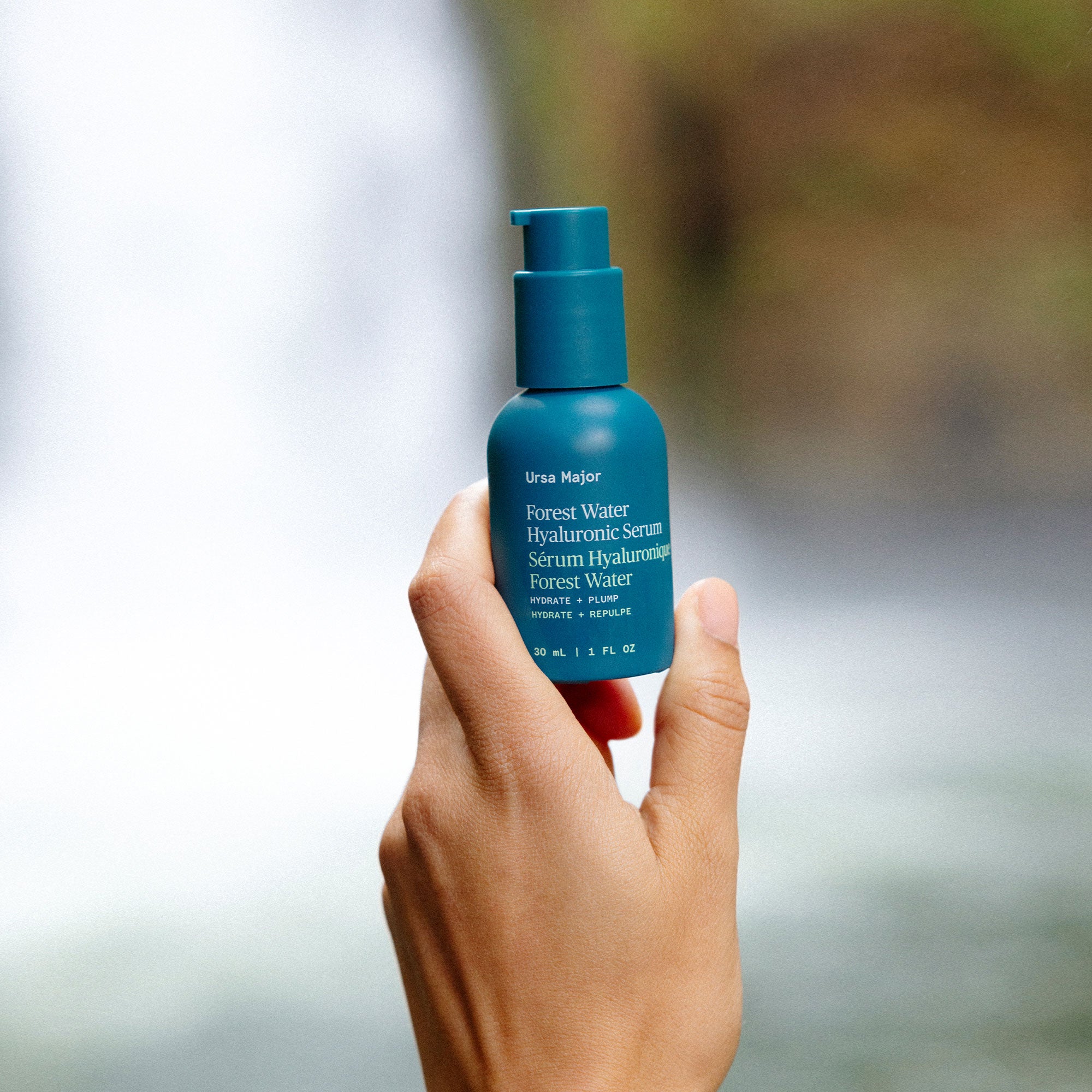 Hand holding a blue bottle of Ursa Major Forest Water Serum against a blurred natural background