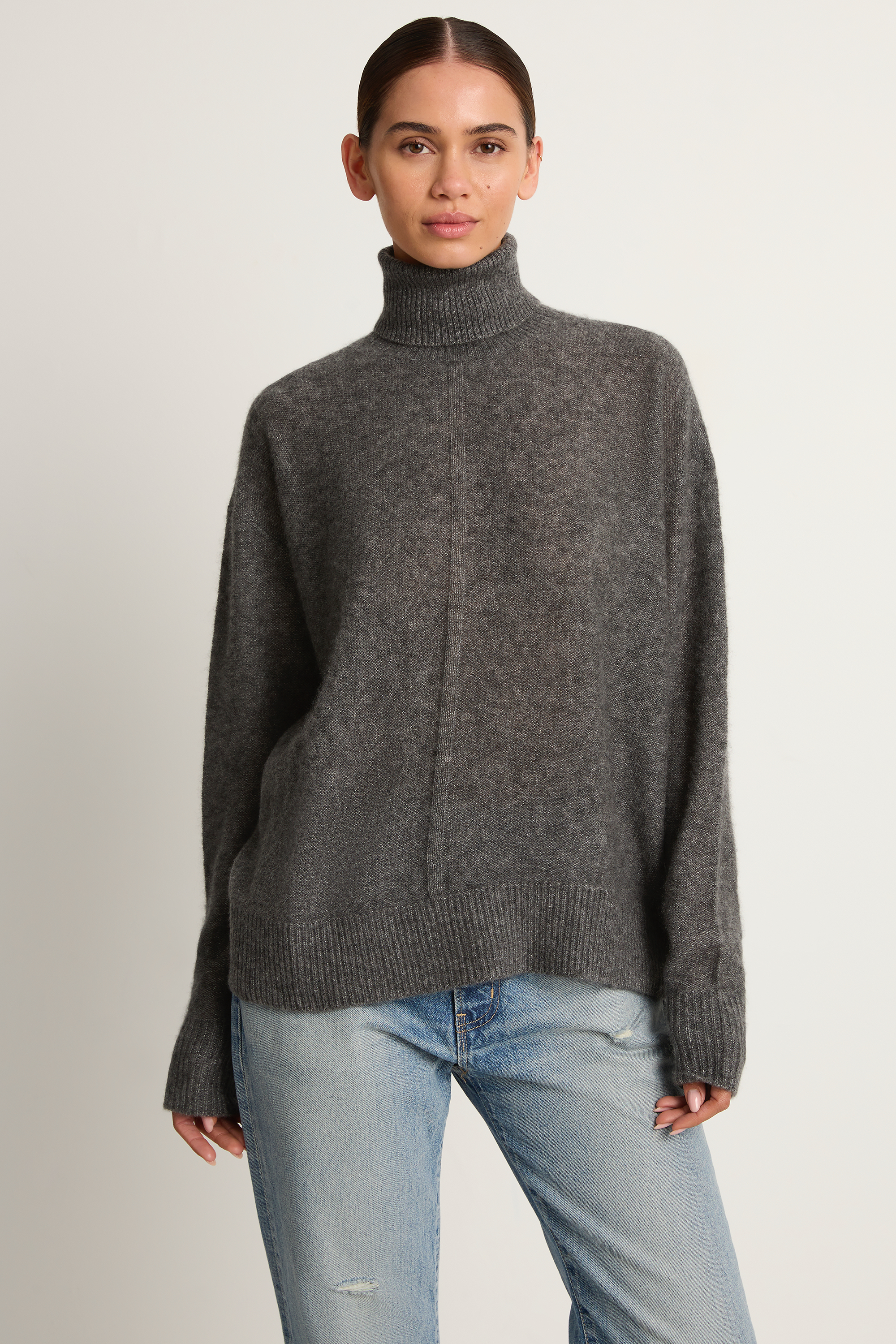 Woman wearing a gray turtleneck sweater and light blue jeans on a plain background