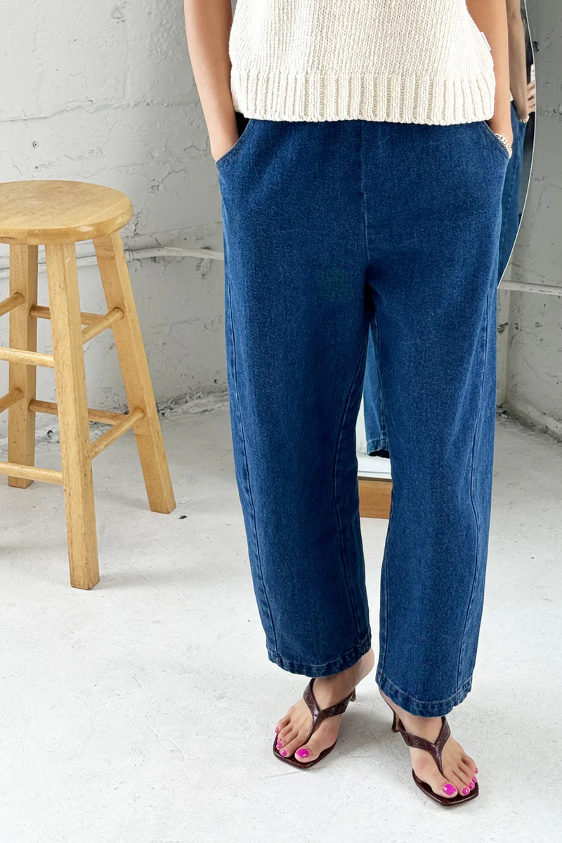 Person wearing blue jeans and a white top in a minimalistic setting with a wooden stool.