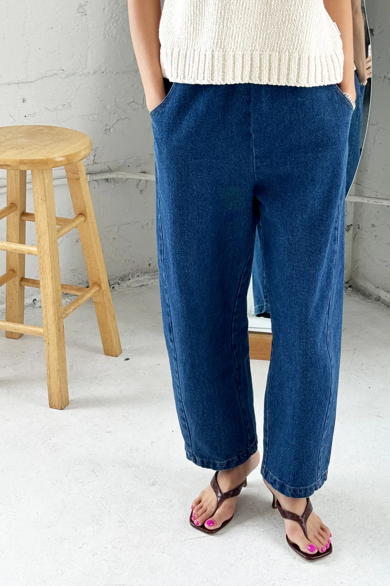 Person wearing blue jeans and a white top in a minimalistic setting with a wooden stool.