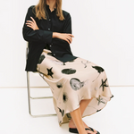 Woman sitting on a chair wearing a black shirt and a patterned skirt with a white background