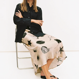 Woman sitting on a chair wearing a black shirt and a patterned skirt with a white background