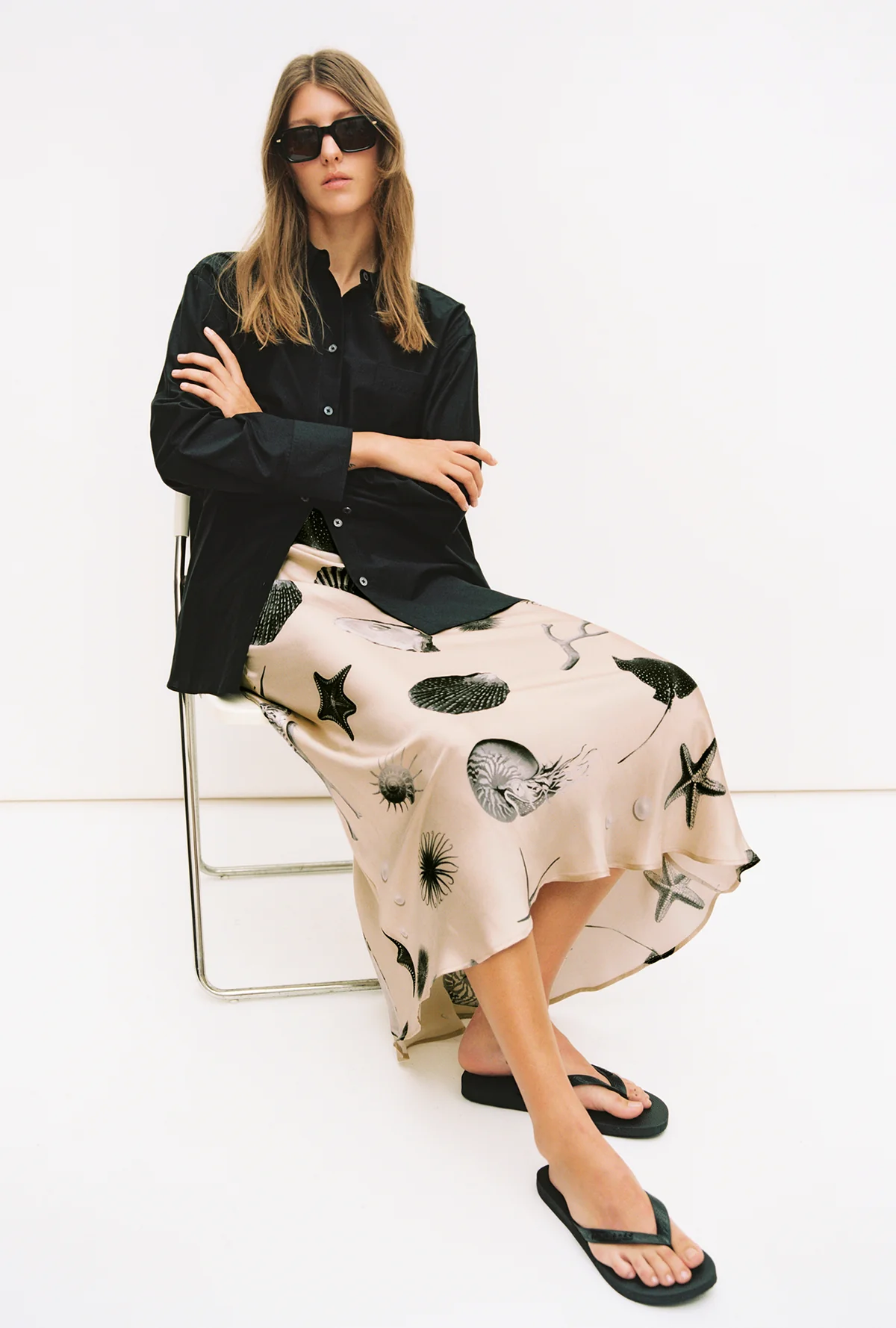 Woman sitting on a chair wearing a black shirt and a patterned skirt with a white background